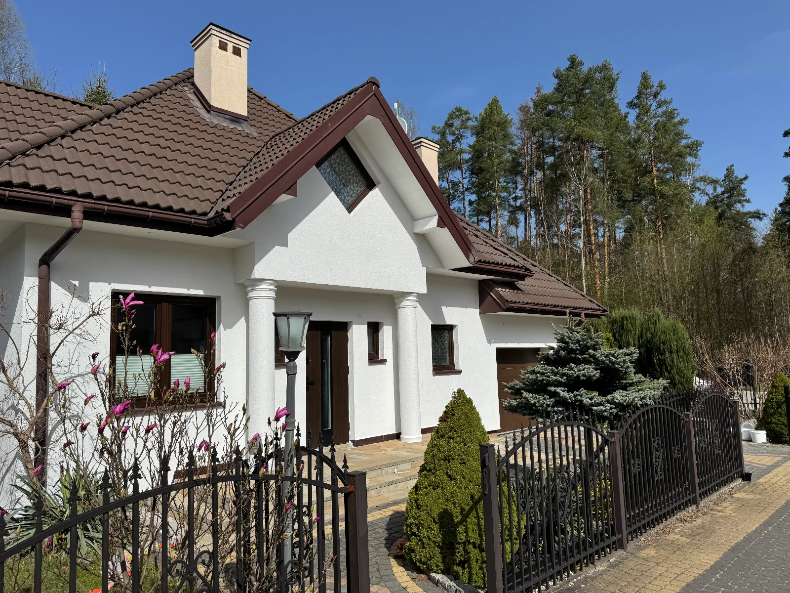 Elewacja domu z białym tynkiem, kolumnowym wejściem, ciemnym dachem i ogrodzeniem metalowym