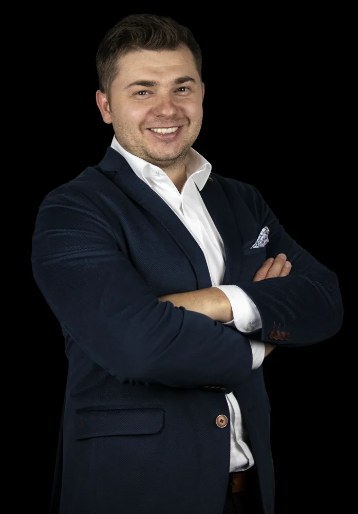 Mężczyzna w eleganckim garniturze i białej koszuli, uśmiechnięty, na czarnym tle