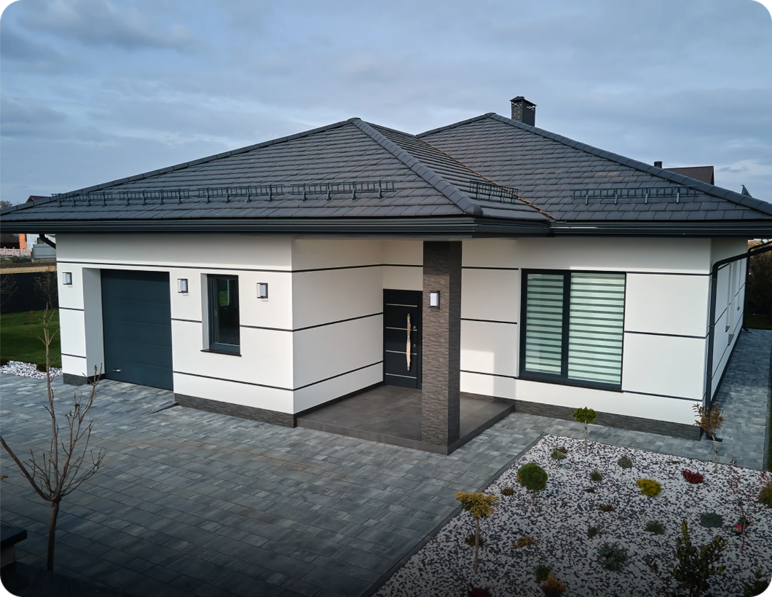 Nowoczesna elewacja domu parterowego z białym tynkiem, czarnymi listwami, płaskim dachem i podjazdem z kostki