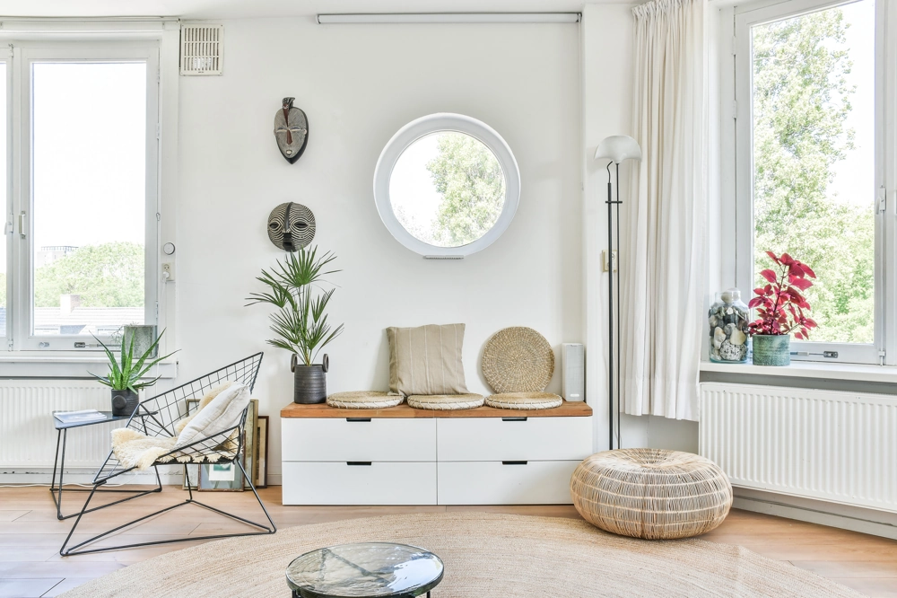 Jasne, minimalistyczne wnętrze salonu z białą ścianą, okrągłym lustrem, ławeczką z poduszkami, roślinami i pufą